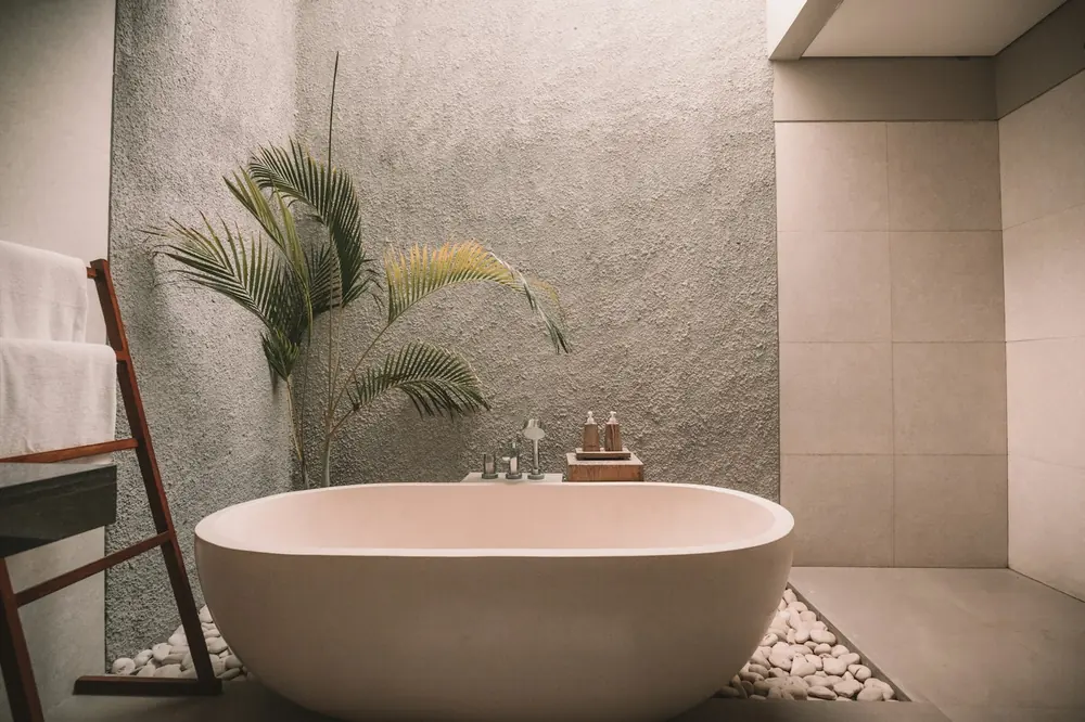 Badkamer met een vrijstaand bad in zandkleurige steen, een muur in ruwe kalkpleister en een palm in strijklicht