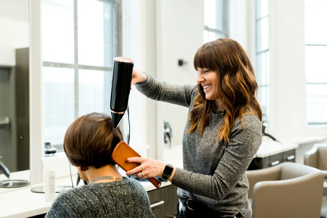 Lotte föhnt lachend het haar van een klant bij het raam van het salon